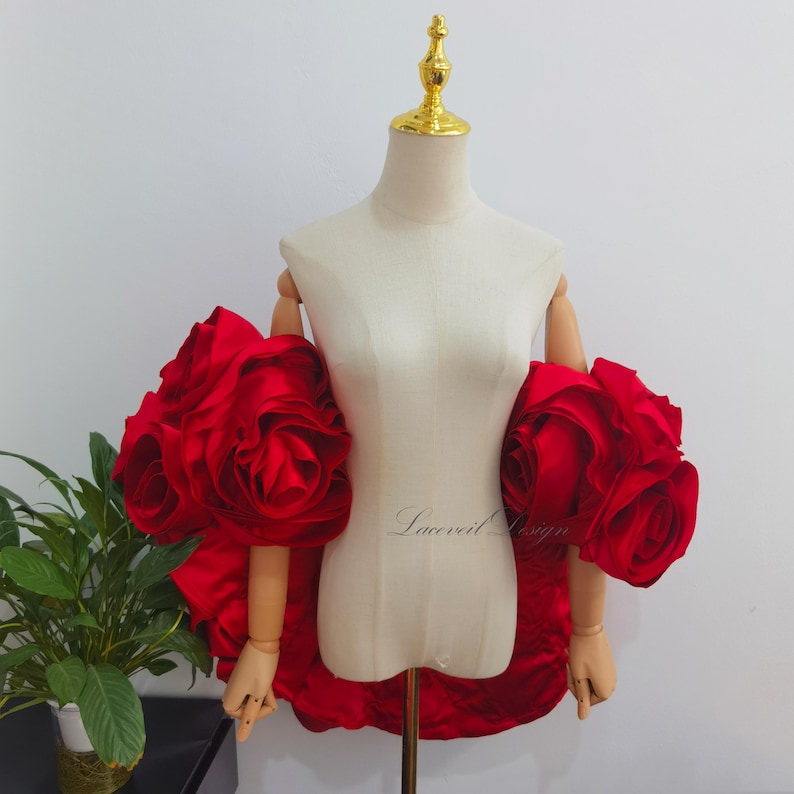K&ouml;nnte beinhalten: Ein rotes Satinkleid mit gro&szlig;en rosenf&ouml;rmigen &Auml;rmeln wird auf einer Schaufensterpuppe pr&auml;sentiert. Die &Auml;rmel sind der Mittelpunkt, mit mehreren Stoffschichten, die das florale Design bilden. Die Schaufensterpuppe hat Holz&auml;rmel und einen goldfarbenen Aufsatz.