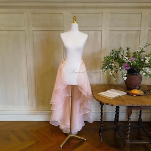 May include: A light pink, high-low skirt with layered ruffles is displayed on a white dress form with a gold stand. The skirt has a flowing, sheer appearance. The dress form has the text "Laceveil Design" printed on it.
