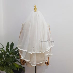 Peut inclure: Un voile de mariée blanc à deux niveaux avec une bordure en ruban de satin. Le voile est fait d'un tissu transparent et léger et est présenté sur un mannequin. Le texte "Laceveil Design" est visible.