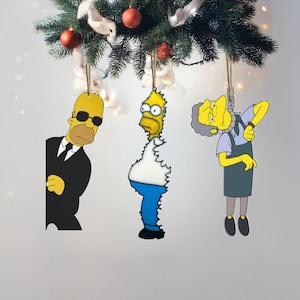 Może przedstawiać: Ozdoby choinkowe z postaciami z kreskówek Homerem Simpsonem, Homerem Simpsonem w czarnym garniturze i Moe Szyslakiem. Ozdoby są żółte, niebieskie, czarne i szare, ze sznurkiem do zawieszenia. Ozdoby wiszą na udekorowanej choince.