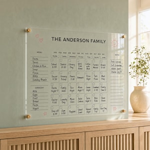 May include: A clear acrylic wall calendar titled The Anderson Family with black printed text. It features a monthly grid with daily checkboxes, a menu section, a grocery list, and a notes column. The board is mounted with four gold hardware standoffs.