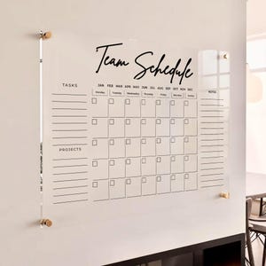 Puede incluir: Un tablero de planificación de equipo de acrílico transparente montado en la pared con texto y líneas de cuadrícula impresas en negro. Incluye secciones para tareas, proyectos y notas, con un calendario mensual, fijado por cuatro soportes metálicos dorados.