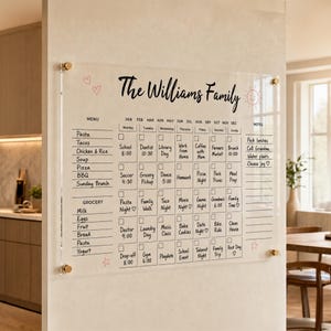Könnte beinhalten: Ein Wandkalender aus klarem Acryl mit der Aufschrift The Williams Family, einem Monatsraster für tägliche Aufgaben, einem Menübereich und einer Notizspalte. Die Tafel ist an jeder Ecke mit goldfarbenen Halterungen an der Wand befestigt.