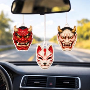 May include: Three hanging car air fresheners suspended from a rearview mirror. The left is a red oni mask with gold horns, the right is a pale oni mask with gold horns, and the center is a white fox mask with red markings and black details.