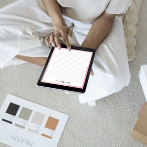 K&ouml;nnte beinhalten: Eine Person sitzt auf einem Teppich und verwendet ein Tablet, um eine To-Do-Liste zu erstellen. Der Tablet-Bildschirm zeigt eine rosa-wei&szlig;e To-Do-Listen-Vorlage. Neben der Person ist eine Farbpalette auf dem Boden sichtbar.