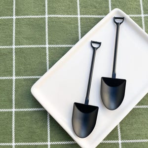May include: Two matte black spoons shaped like miniature shovels with D-shaped handles, resting on a rectangular white ceramic tray. The spoons feature a wide, curved bowl and a long, slender stem, providing a distinct, functional design.
