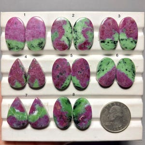 May include: A collection of eight pairs of polished ruby zoisite cabochons in oval and teardrop shapes, arranged in numbered rows on a white display tray next to a United States quarter coin for scale, featuring mottled pink, green, and black patterns.