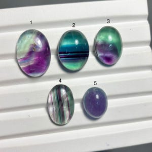 Könnte beinhalten: Fünf ovale Fluorit-Edelsteine in verschiedenen Farben. Die Steine zeigen eine Reihe von Farbtönen, darunter Lila, Grün und Blau, mit durchscheinenden und undurchsichtigen Bereichen. Sie sind auf einer weißen Oberfläche angeordnet.