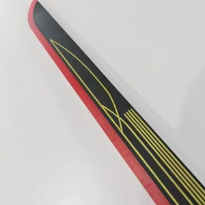 May include: A black and red ruler with yellow geometric designs. The ruler has a red edge with measurement markings and a black center with yellow lines and shapes. The ruler is angled diagonally against a white background.