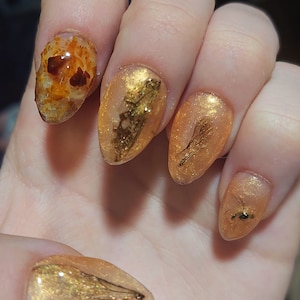 Könnte beinhalten: Eine Hand mit fünf langen, mandelförmigen Nägeln. Die Nägel sind mit einem schimmernden orange-goldenen Lack lackiert. Einige Nägel weisen Blattgold und kleine, dunkelbraune und schwarze Akzente auf, wodurch ein einzigartiges, künstlerisches Design entsteht.