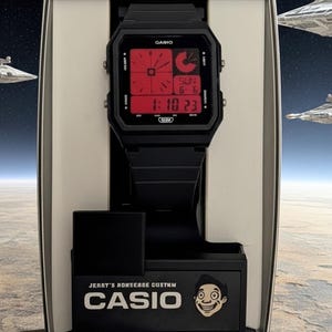 May include: A black digital wristwatch with a square case and a red display screen featuring time, date, and day indicators. The watch is mounted on a black display stand with the text CASIO and a cartoon face graphic printed on the front base.