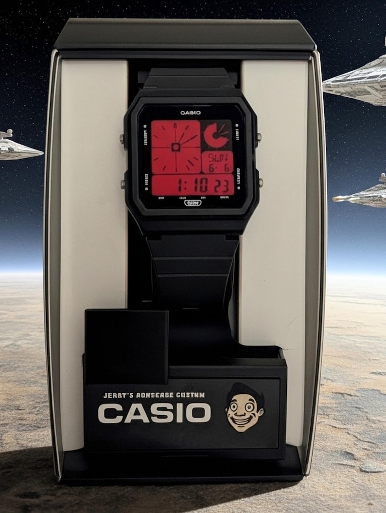 May include: A black digital wristwatch with a square case and a red display screen featuring time, date, and day indicators. The watch is mounted on a black display stand with the text CASIO and a cartoon face graphic printed on the front base.