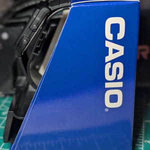 May include: A black digital watch with a rectangular face and a black strap, positioned next to a blue Casio branded display box. The box features the white text CASIO printed vertically on its side, resting on a green mat with inch markings.