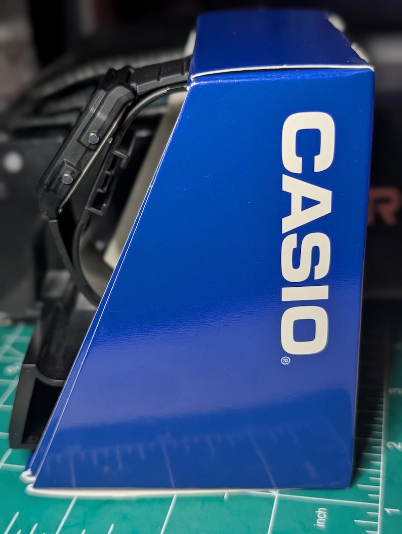May include: A black digital watch with a rectangular face and a black strap, positioned next to a blue Casio branded display box. The box features the white text CASIO printed vertically on its side, resting on a green mat with inch markings.