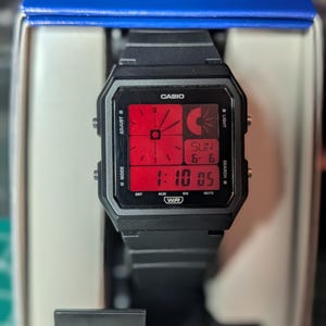May include: A black Casio digital watch with a square case and a red display. The screen shows an analog-style clock face, a digital time display of 1:10 05, and the date SUN 6-6. The watch features side buttons labeled ADJUST, LIGHT, MODE, and SEARCH.