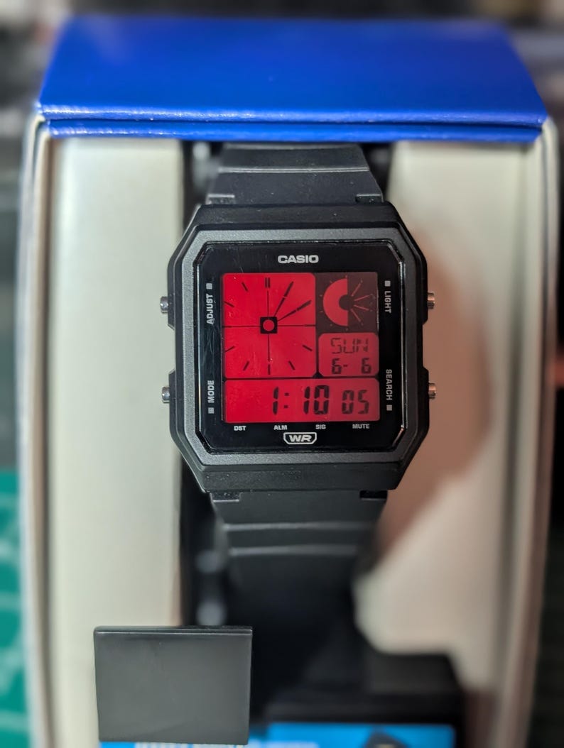 May include: A black Casio digital watch with a square case and a red display. The screen shows an analog-style clock face, a digital time display of 1:10 05, and the date SUN 6-6. The watch features side buttons labeled ADJUST, LIGHT, MODE, and SEARCH.