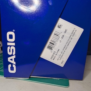 May include: A blue Casio product box with white text. A white label on the side displays the model number LF-20W-1AKC, a barcode, and the text: Not a toy. Not intended for children under 12. Distributed by Casio America, Inc. Dover, NJ. Made in China.