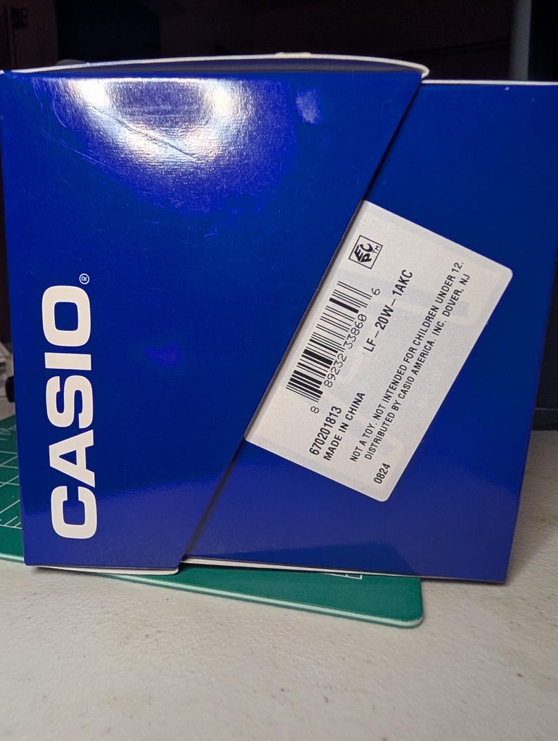 May include: A blue Casio product box with white text. A white label on the side displays the model number LF-20W-1AKC, a barcode, and the text: Not a toy. Not intended for children under 12. Distributed by Casio America, Inc. Dover, NJ. Made in China.