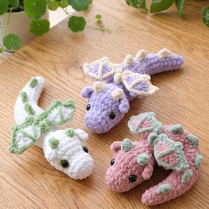 Może przedstawiać: Trzy ręcznie robione szydełkowe smoki w kolorze białym, lawendowym i różowym z zielonymi akcentami. Smoki mają skrzydła i są ułożone na drewnianym stole. Smoki to fantazyjny, ręcznie robiony przedmiot.