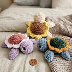 May include: Three handmade crochet turtle plush toys in shades of blue, purple, and beige. Each turtle has a unique shell design, including a sunflower and a flower. A small wooden tag with the words "Handmade with Love" is also visible.