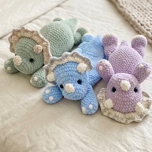 May include: Four handmade crochet dinosaur toys in shades of green, blue, and purple, with white and gray accents. The plush toys are lying on a cream-colored bedspread. Each dinosaur has a unique design, including horns and frills.