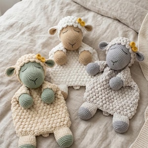 以下が含まれることがあります： クリーム、グリーン、グレーの3色の手編みひつじのおもちゃ。 それぞれのひつじは、質感のある体、閉じられた目、黄色の花のアクセントが付いています。 おもちゃはニュートラルな色合いのベッドカバーの上に置かれており、柔らかく魅力的なシーンを作り出しています。