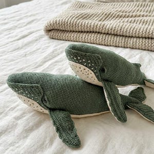 Könnte beinhalten: Zwei handgefertigte, grüne Wal-Plüschtiere aus Häkelarbeit liegen auf einer weißen Bettdecke. Die Wale haben cremefarbene Bäuche und Flossen. Eine gefaltete beige Decke und ein Kissen befinden sich im Hintergrund und schaffen ein gemütliches, maritimes Thema.