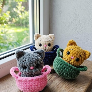 Op de afbeelding: Vier gehaakte kattenspeeltjes in verschillende kleuren: grijs, crème, geel en groen. Elke kat zit in een bijpassend mandje met handvatten. De katten hebben roze neuzen en zwarte ogen. De speeltjes worden op een houten oppervlak gepresenteerd.