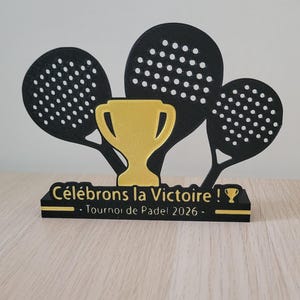 Peut inclure: Trophée noir et or avec trois raquettes de padel et une coupe dorée. La base indique "Célébrons la Victoire !" et "Tournoi de Padel 2026".