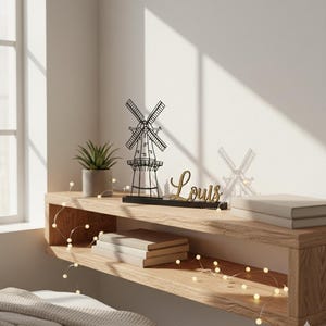 Peut inclure: Une étagère flottante en bois avec une décoration de moulin à vent, une petite plante en pot et le nom "Louis" en lettres dorées. L'étagère contient également des livres et des guirlandes lumineuses. Le mur est blanc.