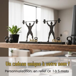 Peut inclure: Silhouettes noires de personnes soulevant des poids sur une étagère en bois. Chaque figurine tient une barre au-dessus de sa tête. Les noms "Julien" et "Clara" sont en lettres dorées. L'étagère contient également une plante en pot, des kettlebells et un tapis de yoga. Texte: "Un cadeau unique à votre nom !"