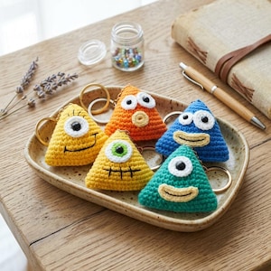 May include: A collection of colorful, handmade crochet monster keychains in yellow, orange, blue, and green. Each triangular keychain features a unique eye and smiling mouth design, attached to a gold-colored keyring. They are arranged on a decorative tray.