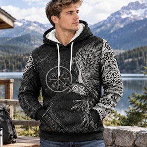 Op de afbeelding: Een zwarte hoodie met een witte sherpa voering, witte trekkoorden en een gedetailleerde grafische print van een raaf, ingewikkelde Keltische knopen en een Vegvisir kompassymbool op de voorzijde en de mouwen met een textuur.