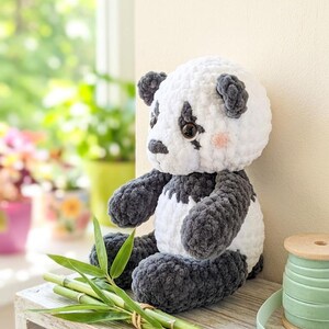 Pode incluir: Um urso panda de crochê feito à mão, branco e cinza, sentado em uma prateleira de madeira. O panda tem olhos pretos e bochechas rosadas. Caules de bambu e um carretel de fita verde completam a cena.