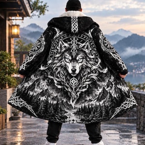 May include: A black and white hooded cloak featuring a large wolf head graphic, intricate knotwork patterns, and a landscape of mountains and pine trees. The garment has a white sherpa fleece lining visible at the collar and hood, with a long, flowing hem.