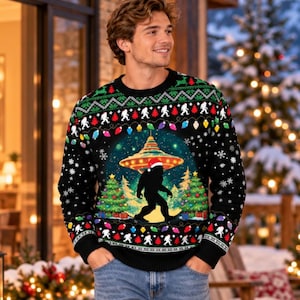Pode incluir: Um suéter preto de manga comprida e gola redonda com uma estampa gráfica da silhueta do Bigfoot usando um gorro de Papai Noel, em pé entre duas árvores de Natal decoradas sob um OVNI brilhante, com uma borda de luzes e flocos de neve.