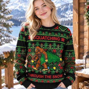 May include: A black long-sleeve crewneck sweater featuring a festive holiday pattern in red and green. The design includes a graphic of a brown Bigfoot carrying a green Christmas tree, with the text SQUATCHING THROUGH THE SNOW printed in red.