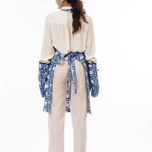 May include: A blue and white floral print apron with long sleeves and two pockets. The apron is tied at the waist with a blue ribbon.