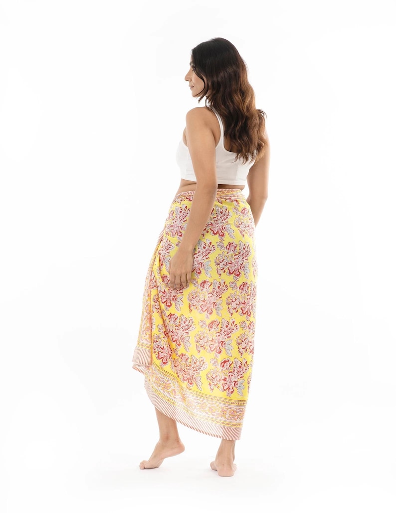 May include: A bright yellow sarong featuring a repeating floral pattern in shades of red and white. The fabric is wrapped around the waist, extending to the lower calf, and is finished with a striped border along the bottom edge and side.
