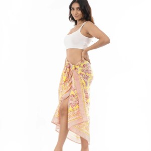 May include: A yellow sarong featuring a vibrant floral pattern in shades of pink, orange, and white. The fabric is tied at the waist, creating a draped silhouette with a side slit. The sarong is paired with a simple white sleeveless crop top.