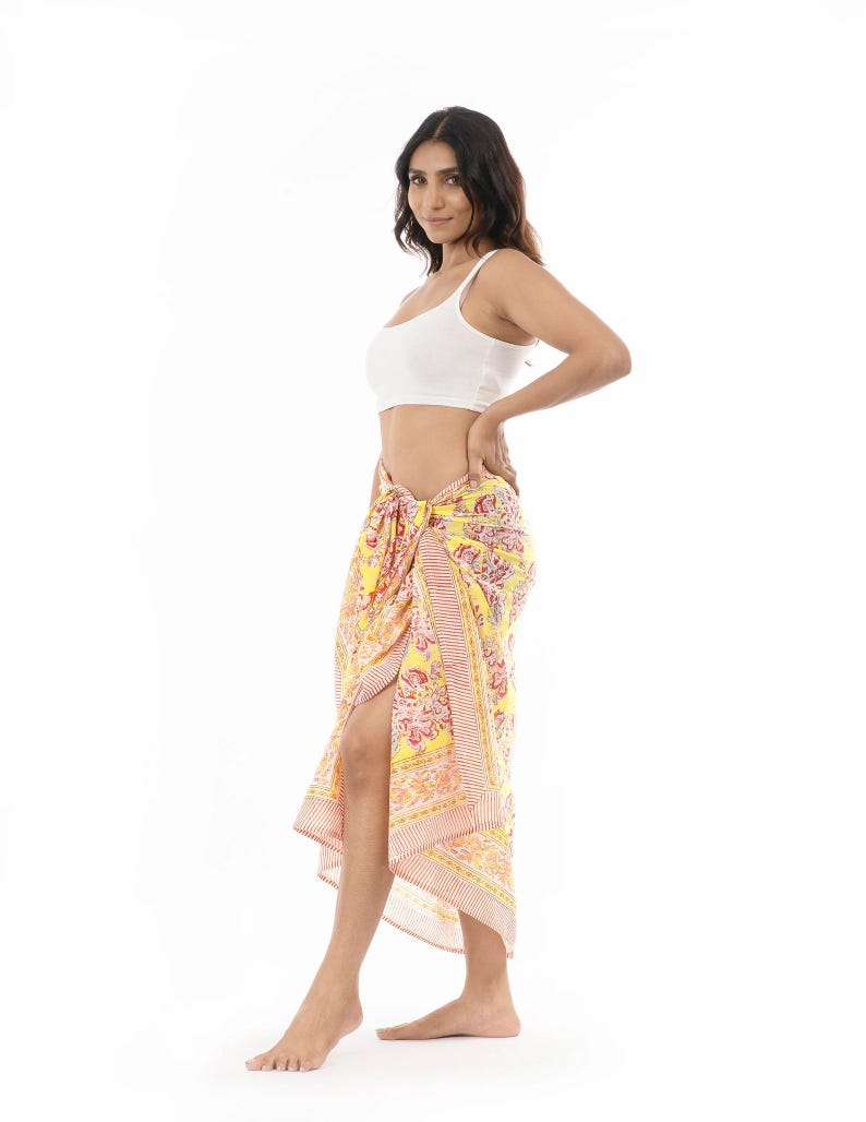 May include: A yellow sarong featuring a vibrant floral pattern in shades of pink, orange, and white. The fabric is tied at the waist, creating a draped silhouette with a side slit. The sarong is paired with a simple white sleeveless crop top.