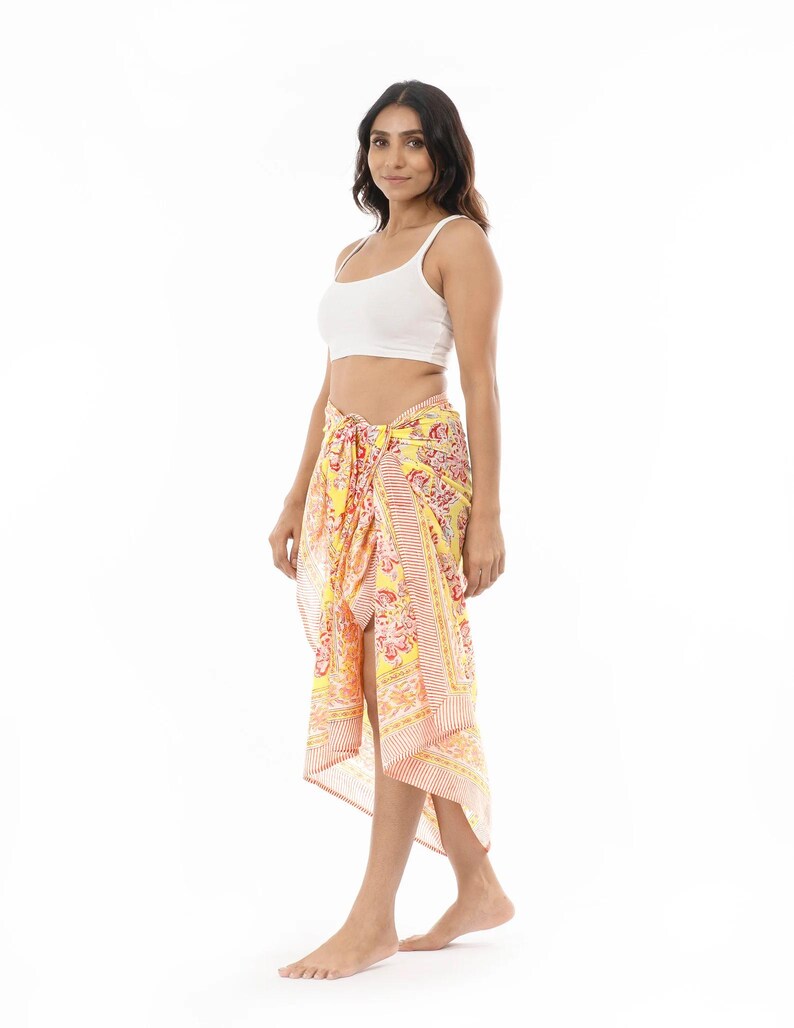 May include: A yellow sarong featuring a vibrant floral pattern in shades of pink, orange, and white. The fabric is tied at the waist, creating a draped silhouette with a side slit. The design includes a striped border detail along the edges.