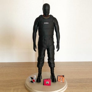 May include: Black action figure wearing a black vest with the word "DONDA" printed on it. The figure is standing on a round platform with four miniature CDs.