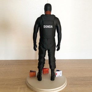 May include: Black figurine of a person wearing a black vest with the word "DONDA" printed on it. The figurine is standing on a round base with three CDs on it.