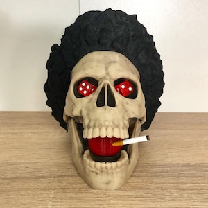 Peut inclure: Figurine de crâne fantaisie avec des dés rouges à la place des yeux et une cigarette dans la bouche. Le crâne a une afro noire et une boule rouge dans la bouche. Le crâne est en matériau beige et repose sur une surface en bois.