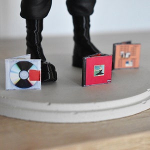 May include: Three miniature vinyl records on a white platform. The records are a red album with a black and white image of a woman in a black dress, a brown album with a white and black image of a building, and a clear CD with a red sticker.