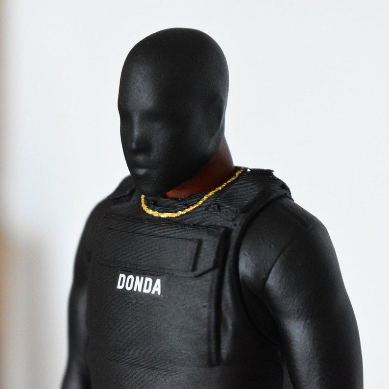 May include: Black and gold figurine wearing a black vest with the word "DONDA" printed on it.