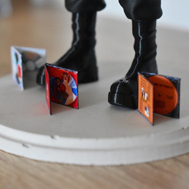 May include: A black figure with boots standing on a white platform. There are four miniature vinyl records with colourful covers on the platform.