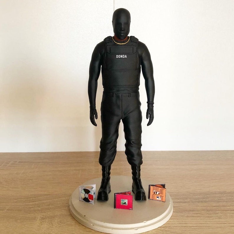 May include: Black action figure wearing a black vest with the word "DONDA" printed on it. The figure is standing on a round platform with four miniature CDs.
