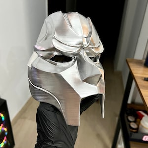 May include: A silver-colored mask with a textured surface and a sharp, angular design. The mask has a raised, geometric pattern and a pointed chin.